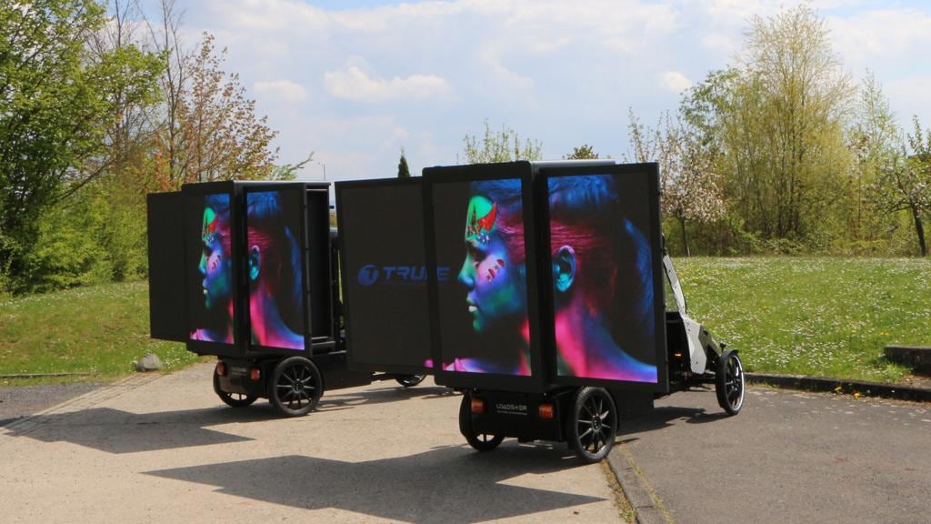 Two TRUE dvLED trailers parked outdoors on a path, both screens showing a colour portrait in daylight