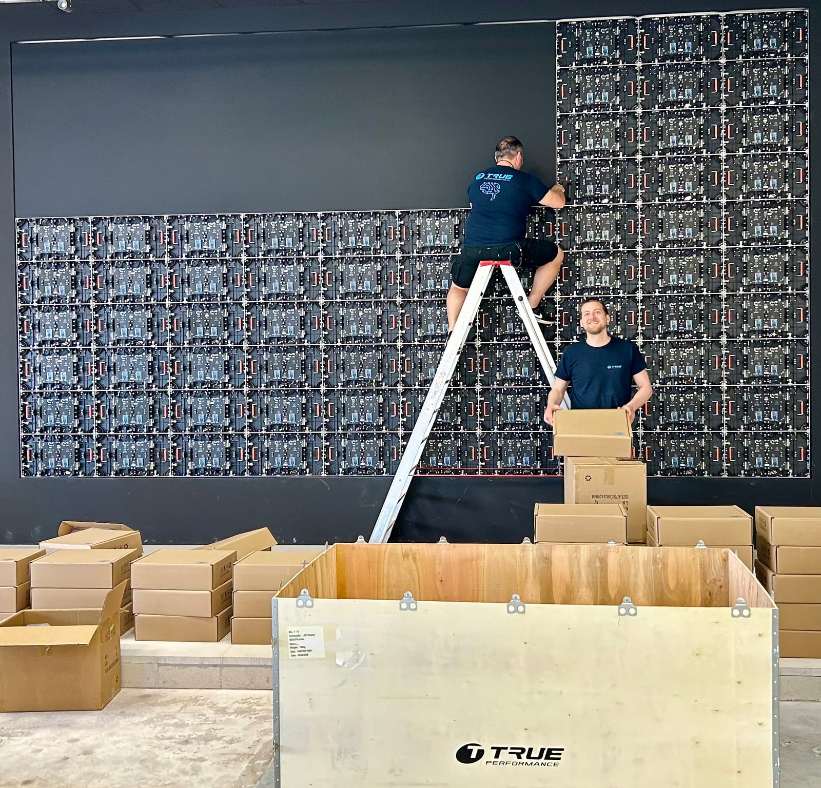 Two TRUE technicians mounting dvLED panels from a ladder, the TRUE transport crate and boxed panels in front of the dvLED wall